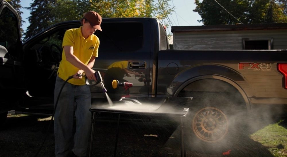 A detailer in a yellow polo spraying a truck's floor mat on a folding table in a driveway
